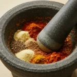 A small bowl filled with homemade burrito seasoning, surrounded by individual spices like chili powder, cumin, and paprika on a wooden table.