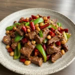 A sizzling plate of Kung Pao Beef with tender slices of beef, colorful diced bell peppers, and roasted peanuts, coated in a glossy savory-spicy sauce. Served over steamed rice and garnished with green onions.