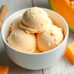 A bowl of refreshing melon ice cream with a vibrant orange color, garnished with fresh melon slices and mint leaves.