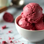 A bowl of creamy red velvet ice cream with a vibrant red color, topped with crumbled red velvet cake and a drizzle of white chocolate.